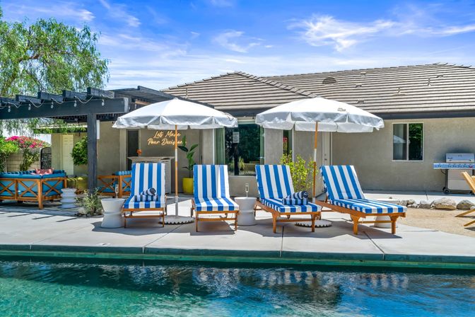 Resort-style backyard pool deck with four blue-and-white striped lounge chairs under white umbrellas, pergola seating area, neon wall sign and stainless barbecue grill.