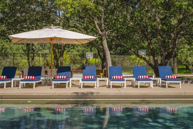 Sunny outdoor poolside scene: row of white loungers with blue cushions and red-and-white striped towels under a large beige umbrella, reflected in the water with leafy trees and a basketball hoop in the background.