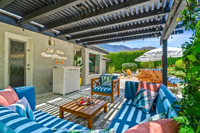 Sunny backyard patio under a slatted pergola with string lights, bright blue cushioned lounge set and wooden coffee table with cocktails, striped poolside loungers and white umbrellas, manicured hedges and mountain backdrop.