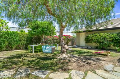 Sunny suburban backyard with a large shady tree and round hanging blue-cushioned swing, white dining table with lime-green chairs set for al fresco dining, cornhole boards on the lawn, stone path and flowering hedges by a house.