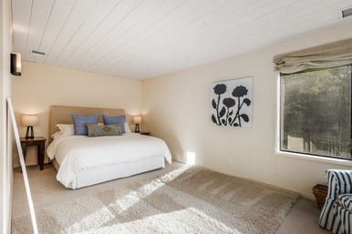 Bright airy master bedroom with white shiplap ceiling, king bed with blue accent pillows, wooden nightstands with lamps, plush beige rug and a large window overlooking trees