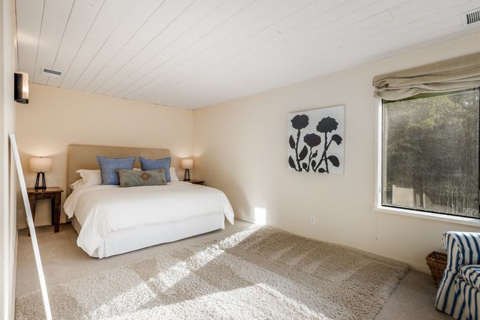 Bright airy master bedroom with white shiplap ceiling, king bed with blue accent pillows, wooden nightstands with lamps, plush beige rug and a large window overlooking trees