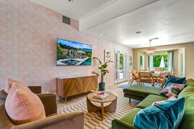 Sunlit open-plan living and dining room with pink geometric accent wall, green velvet sectional, blush-pink pillows, wooden media console, round coffee table and glass doors opening to a leafy backyard patio.