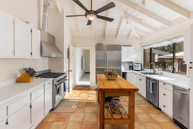 Sunlit modern kitchen with white vaulted beams, wooden butcher-block island, stainless steel range and refrigerator, large window over sink and terracotta tile floor