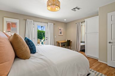 Modern sunlit bedroom with white bedding, blush and navy throw pillows, French doors opening to a sunny patio with lounge chairs and palm trees, small wood desk and hardwood floors