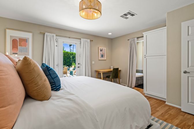 Modern sunlit bedroom with white bedding, blush and navy throw pillows, French doors opening to a sunny patio with lounge chairs and palm trees, small wood desk and hardwood floors