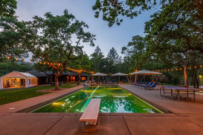 Twilight outdoor pool at a wooded country retreat—diving board over green-lit water, twinkling string lights draped between trees, poolside umbrellas and blue lounge chairs with a tented seating area.