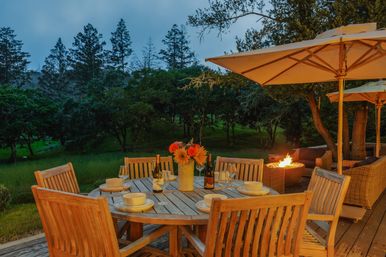 Cozy evening outdoor dining on a wooden deck: round table set for six with wine bottles, glasses and a vase of flowers, large patio umbrellas and a glowing fire pit overlooking a tree‑covered hillside.