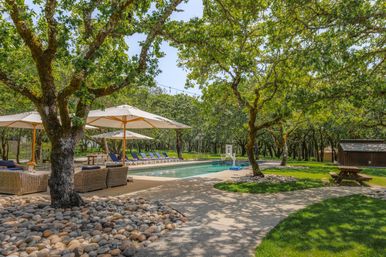 Sunlit outdoor pool framed by mature oak trees, wicker lounge seating and large umbrellas, a row of blue chaise lounges, stone-bordered patio and picnic table in a shaded wooded yard.