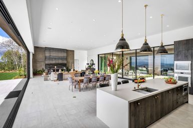 Bright modern open‑concept kitchen and dining great room with a large white island and brass faucet, dark wood cabinets, three black pendant lights, long wooden dining table with upholstered chairs, tall floral centerpiece, and floor‑to‑ceiling glass walls opening to a patio with ocean view and lush lawn.