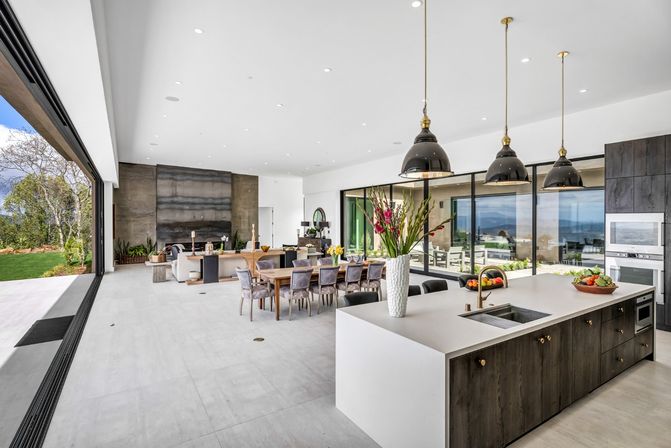 Bright modern open‑concept kitchen and dining great room with a large white island and brass faucet, dark wood cabinets, three black pendant lights, long wooden dining table with upholstered chairs, tall floral centerpiece, and floor‑to‑ceiling glass walls opening to a patio with ocean view and lush lawn.