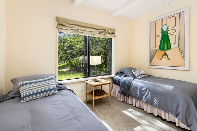 Sunlit twin bedroom with gray bedding and striped pillows, wooden nightstand and lamp between beds, large window with beige roman shade overlooking green trees, and wall art of a green dress.