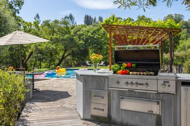 Sunny backyard patio with built-in stainless-steel outdoor kitchen grill topped with colorful peppers and corn, vine-covered wooden pergola, and a pool with a yellow duck float and striped umbrella against a lush hillside backdrop