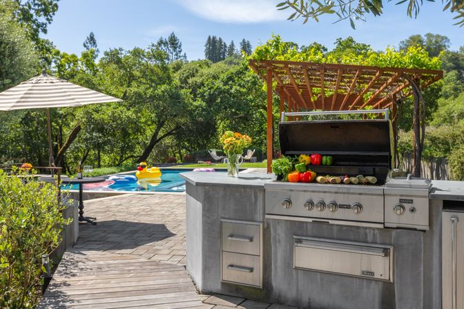 Sunny backyard patio with built-in stainless-steel outdoor kitchen grill topped with colorful peppers and corn, vine-covered wooden pergola, and a pool with a yellow duck float and striped umbrella against a lush hillside backdrop