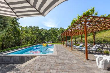 Sunny hillside backyard oasis with rectangular swimming pool and colorful inflatable floats, striped patio umbrella, wooden pergola with climbing vines, cushioned lounge chairs on a paver patio surrounded by oak trees.