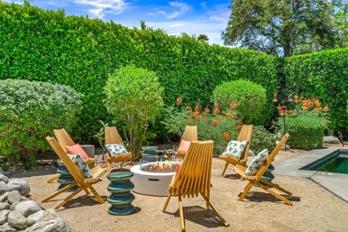 Sunny backyard patio with a round fire pit surrounded by six wooden slatted lounge chairs with tropical pillows, green ceramic side tables, sandy gravel ground, tall manicured hedges, orange flowering shrubs and a nearby swimming pool under a blue sky.