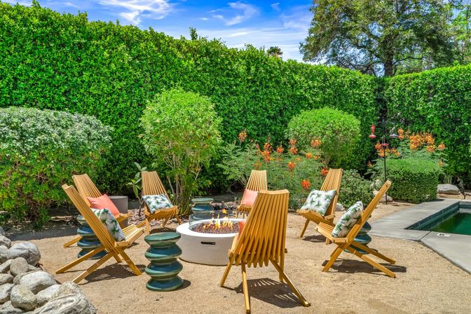 Sunny backyard patio with a round fire pit surrounded by six wooden slatted lounge chairs with tropical pillows, green ceramic side tables, sandy gravel ground, tall manicured hedges, orange flowering shrubs and a nearby swimming pool under a blue sky.
