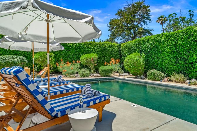 Sunny residential backyard pool with blue-green water, wooden lounge chairs featuring blue-and-white striped cushions and rolled towels under large white umbrellas, set on a concrete deck and surrounded by manicured hedges and flowering shrubs.