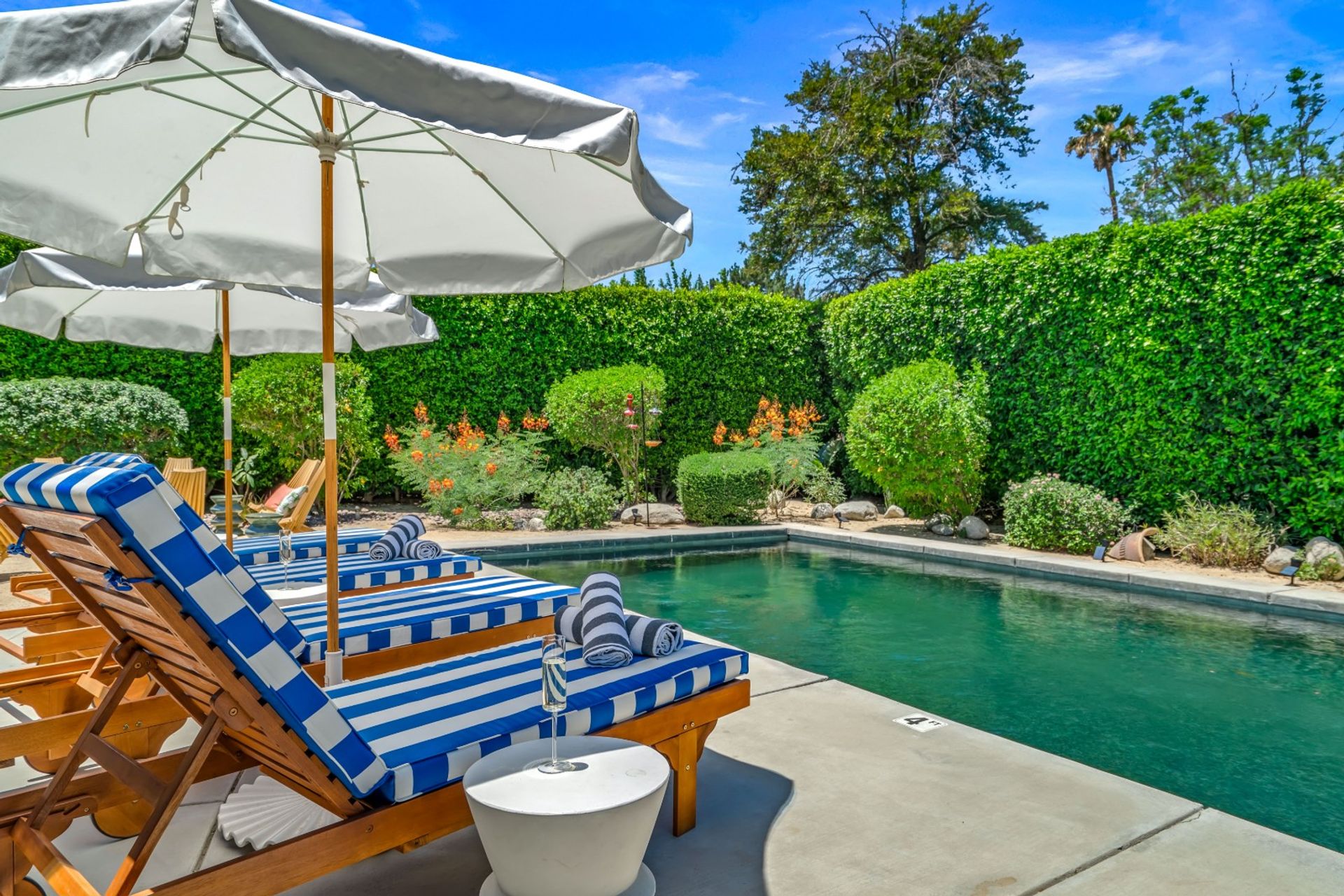Sunny residential backyard pool with blue-green water, wooden lounge chairs featuring blue-and-white striped cushions and rolled towels under large white umbrellas, set on a concrete deck and surrounded by manicured hedges and flowering shrubs.