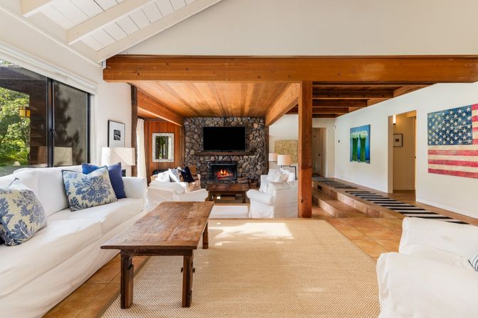 Cozy sunlit living room with vaulted white ceiling and exposed wood beams, stone fireplace with TV, white slipcovered sofas, rustic wooden coffee table, large window overlooking a green yard, and American flag wall art.