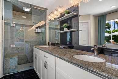 Spa-like sunlit bathroom with a granite double-sink vanity, frameless glass walk-in shower, floating stone shelves with potted plants, and sparkling crystal vanity lights overlooking a sunny backyard.