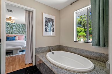 Oval soaking tub set in gray granite surround in a bright bathroom with a window and green curtain overlooking a lush garden and patio chairs; doorway opens to a bedroom with tropical leaf wallpaper, pastel pillows, bench and rattan ceiling light.