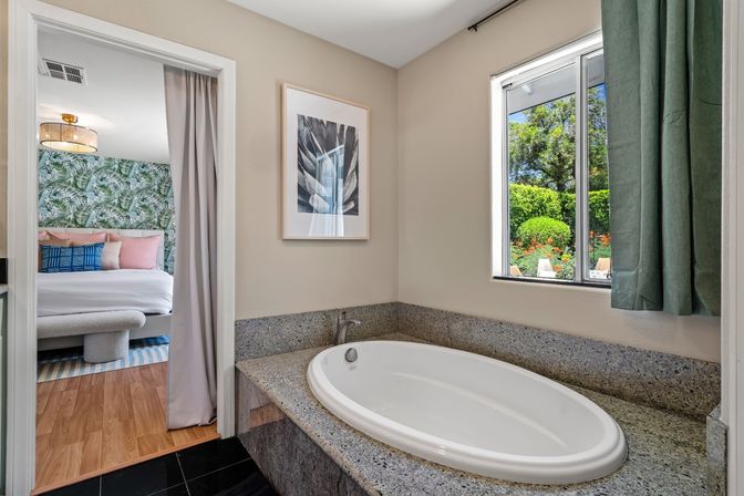 Oval soaking tub set in gray granite surround in a bright bathroom with a window and green curtain overlooking a lush garden and patio chairs; doorway opens to a bedroom with tropical leaf wallpaper, pastel pillows, bench and rattan ceiling light.