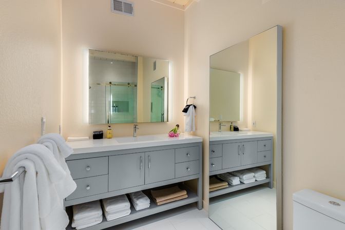 Bright modern bathroom with gray double-sink vanity, illuminated mirrors, neatly folded white towels on open shelves, glass shower reflected in the mirror, and soft beige walls — spa-like and clean.