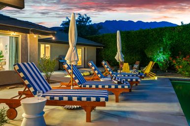 Inviting evening poolside patio with blue-and-white striped lounge chairs, closed umbrellas, warm house lights and a mountain silhouette at sunset