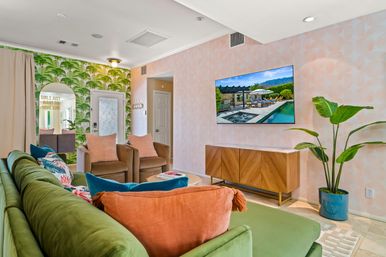 Bright tropical-themed living room with green velvet sofa, coral and teal pillows, palm-leaf wallpaper, two tan armchairs, wall-mounted TV above a mid-century wooden console, and a potted bird-of-paradise plant.