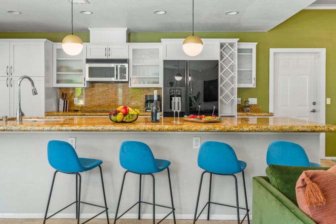 Contemporary open-concept kitchen with green accent wall, granite island topped with fruit bowl, wine bottle and glasses, three blue bar stools, pendant lights and stainless appliances.