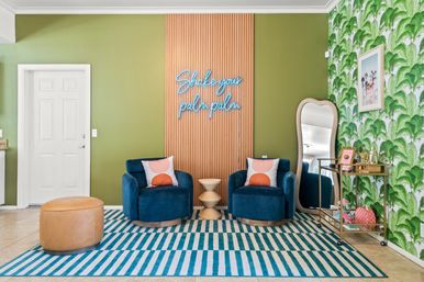 Playful modern tropical lounge with green palm wallpaper, neon 'Shake your palm palm' sign, two blue velvet chairs, striped rug, leather pouf and gold bar cart.