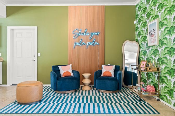 Playful modern tropical lounge with green palm wallpaper, neon 'Shake your palm palm' sign, two blue velvet chairs, striped rug, leather pouf and gold bar cart.