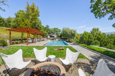 Sunlit hilltop backyard with rectangular swimming pool, wooden pergola with lounge seating, fire pit and cornhole boards overlooking a green wooded hillside.