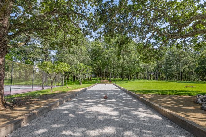 Shaded outdoor bocce ball court with gravel lane and wooden borders, bocce balls clustered in the center, bench at the far end, sun-dappled oak tree canopy, grassy clearing and a fenced tennis court to the left.