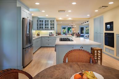 Bright open-concept kitchen with light blue cabinetry, white island with gas cooktop, stainless steel refrigerator, hardwood floors, wicker dining chairs and a fruit bowl on a round table, chandelier over dining area and glass doors to the backyard.