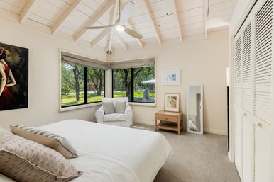 Bright airy bedroom with white vaulted ceiling and ceiling fan, corner windows overlooking a lush green yard, cozy armchair, white bedding and wooden bedside table