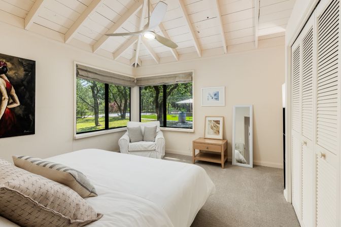 Bright airy bedroom with white vaulted ceiling and ceiling fan, corner windows overlooking a lush green yard, cozy armchair, white bedding and wooden bedside table