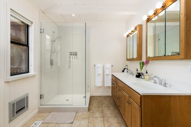 Bright modern farmhouse bathroom with frameless glass walk-in shower, white subway tile, double-sink white countertop and wood vanity, illuminated vanity mirrors with globe bulbs and beige tile floor