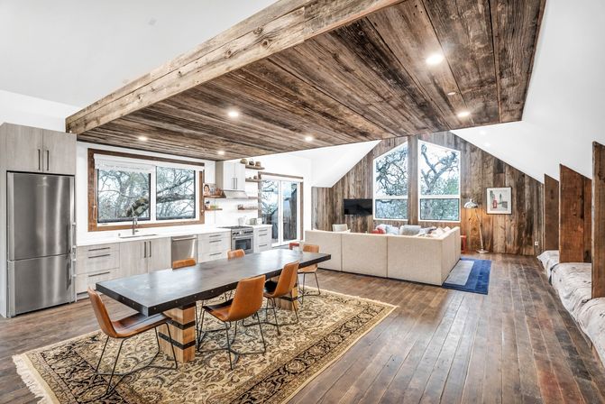 Open-concept rustic-modern kitchen and living room with reclaimed wood ceiling and walls, large windows with tree views, long dining table with leather chairs, stainless fridge and neutral sectional on hardwood floors.