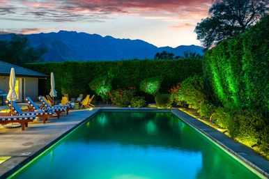 Evening poolside scene: illuminated rectangular swimming pool with striped lounge chairs and umbrellas on a patio, landscaped hedges and lit shrubs, and a colorful sunset over distant mountains.