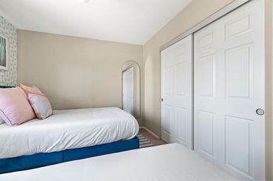 Compact bright bedroom with twin beds, white bedding and pink pillows, blue bed base, geometric accent wall, arched wall mirror and white sliding closet doors