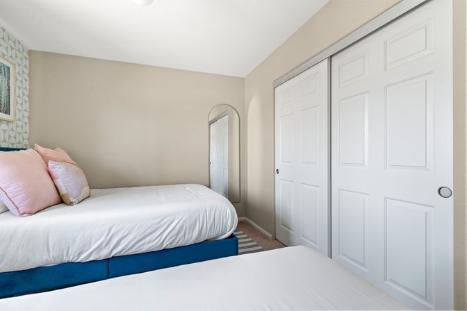 Compact bright bedroom with twin beds, white bedding and pink pillows, blue bed base, geometric accent wall, arched wall mirror and white sliding closet doors