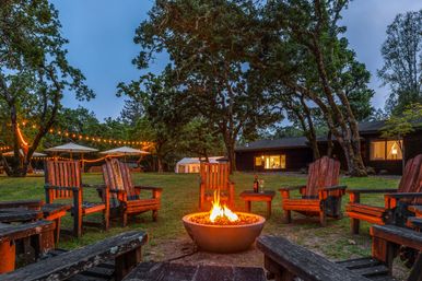 Dusk woodland retreat with a glowing outdoor fire pit surrounded by weathered wooden Adirondack chairs, string lights draped between oak trees, patio umbrellas, and a rustic cabin in the background.