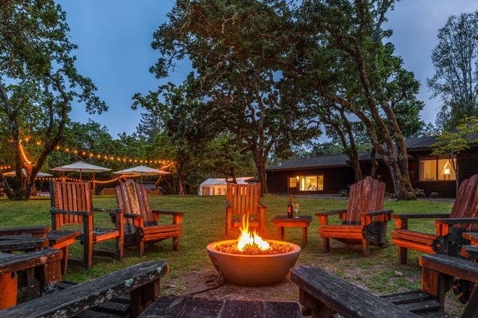 Dusk woodland retreat with a glowing outdoor fire pit surrounded by weathered wooden Adirondack chairs, string lights draped between oak trees, patio umbrellas, and a rustic cabin in the background.