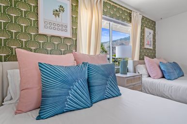 Bright twin bedroom with green patterned wallpaper, pink and teal fan-print pillows, cream curtains and a bedside lamp, window framing palm trees and mountain view