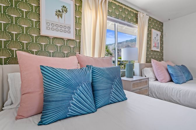 Bright twin bedroom with green patterned wallpaper, pink and teal fan-print pillows, cream curtains and a bedside lamp, window framing palm trees and mountain view