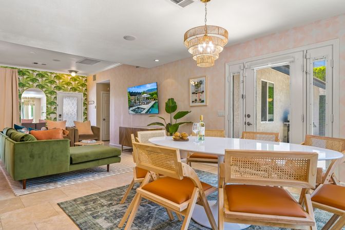 Bright open-plan living and dining room with green velvet sectional, tropical palm wallpaper, round white dining table, cane-back chairs, gold tiered chandelier, and glass French doors to a patio.