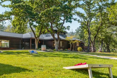 Rustic wooden house nestled among mature oak trees with a sunny green lawn, string lights, wine-barrel planter and cornhole boards for outdoor fun