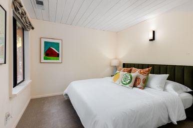 Cozy modern bedroom with large white bed, olive-green upholstered headboard, citrus-print throw pillows, framed colorful barn artwork, bedside lamp, wall sconce and window